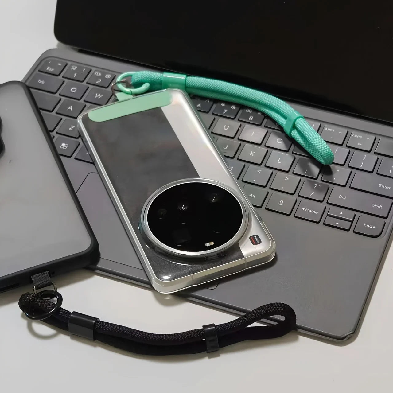 Transparent phone case with green strap on laptop keyboard, nearby black phone case