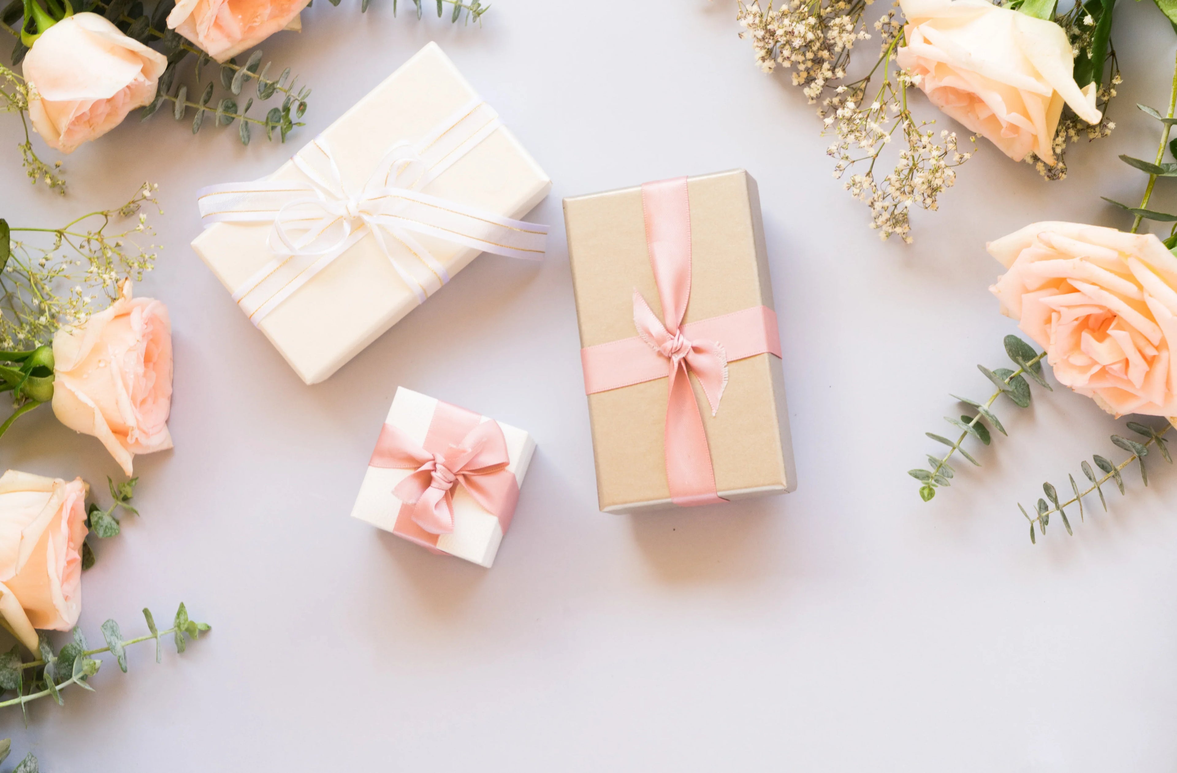 Gift boxes with pink and white ribbons surrounded by peach roses and greenery